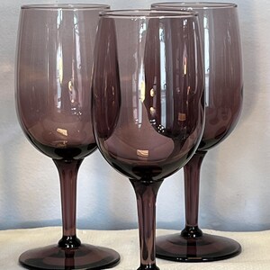 May include: Three stemmed wine glasses with a deep purple hue. The glasses have a classic design with a rounded bowl, a slender stem, and a circular base. The color is consistent throughout the glass, creating a sophisticated look.