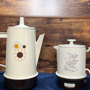 May include: Two vintage electric coffee pots. The larger pot is off-white with a floral design. The smaller pot is also off-white with a coffee bean and leaf design. Both pots have brown bases and white handles.