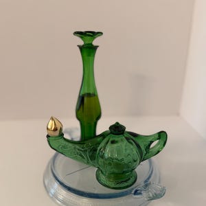 May include: A green glass genie lamp with a gold-toned metal tip and a matching green glass bottle with a tall, slender neck and a stopper, both resting on a clear, circular glass base with a textured edge and a small handle.