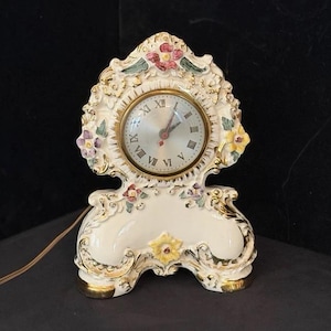 May include: A cream-colored ceramic mantel clock featuring gold-painted scrollwork and hand-painted floral accents in pink, yellow, and green. The clock face has a metallic silver finish with black Roman numerals and two thin, dark-colored hands.