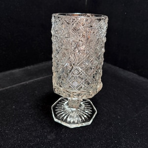 May include: A clear, vintage-style glass with a diamond-cut pattern. The glass has a hexagonal base and a detailed, textured design. The stem is short, and the glass is designed to hold a small amount of liquid.