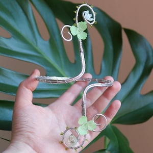 May include: A silver-toned wire ear cuff featuring two spiraling ends decorated with pale green translucent flower-shaped accents and small olive-coloured beads. The metal wire is wrapped around the structure to create a botanical-inspired design.