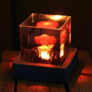 Puede incluir: Un cubo iluminado único con un diseño de explosión en miniatura. La obra de arte está encerrada en resina transparente, con un brillo naranja cálido. El cubo se asienta sobre una base de madera oscura, creando un contraste visual llamativo.
