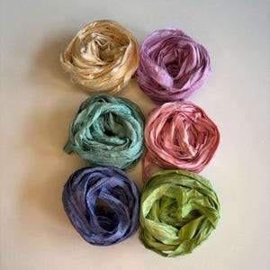 May include: Six circular bundles of frayed-edge silk ribbon arranged in two columns. The colors include cream, lavender, teal, dusty rose, deep blue, and lime green. Each ribbon features a crinkled, textured surface with visible loose threads.