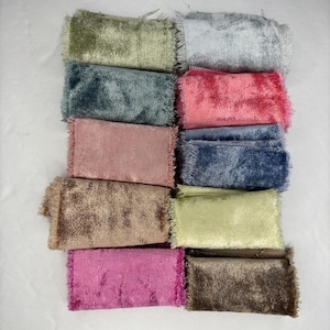 May include: A collection of rectangular fabric swatches in various colors, including green, blue, pink, and brown. Each swatch has a fringed edge, and the fabric appears to be velvet or a similar plush material. The swatches are arranged closely together.