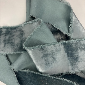 May include: Close-up of a teal velvet ribbon with frayed edges. The ribbon is folded and layered, showcasing its soft texture and color variations. The fabric has a slight sheen, adding depth to the teal hue.
