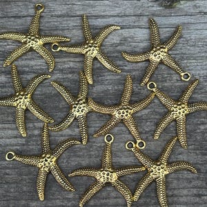 May include: Nine gold-colored starfish charms with textured surfaces and small loops for attaching to jewelry or other crafts. The starfish are arranged on a weathered wooden surface.