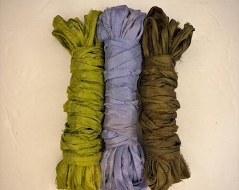 May include: Three bundles of fabric ribbon in shades of green, lavender, and olive. The ribbons are tightly wrapped, with a frayed edge at the bottom. The background is a plain, off-white surface.