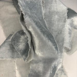 May include: Close-up of several strips of light blue velvet fabric with frayed edges. The fabric has a soft, textured appearance and a subtle sheen. The strips are layered and folded, creating depth and dimension.