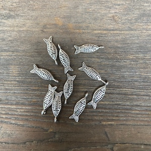 May include: A collection of silver-toned fish charms with detailed designs. The charms have a loop at the top for attaching to jewelry or other crafts.
