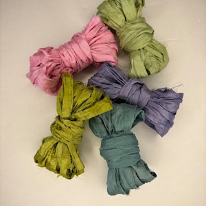 May include: Five bundles of colorful silk ribbon in shades of pink, green, blue, and purple. The ribbons are knotted and have a textured, frayed appearance, suggesting a handmade or artisanal quality. Ideal for crafting or decorative purposes.