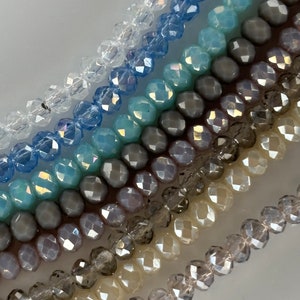 Puede incluir: Primer plano de una variedad de cuentas de vidrio facetadas en tonos de transparente, azul claro, gris y beige. Las cuentas están dispuestas en filas, creando un efecto degradado.