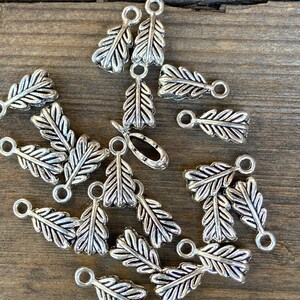 May include: A collection of silver-toned metal leaf-shaped charms with a loop at the top. The charms are scattered on a wooden surface.