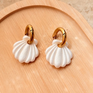 May include: A pair of hoop earrings featuring a polished gold-tone metal hoop and a white, textured seashell-shaped pendant. The shell charms have a subtle pearlescent finish and are suspended from the circular metal hoops.