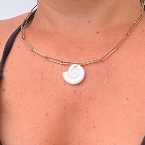 Può includere: Una collana a catena color oro con maglie rettangolari allungate e un singolo ciondolo a forma di conchiglia bianca a spirale. Il ciondolo è sospeso al centro della catena metallica, composta da maglie rettangolari unite in un anello.