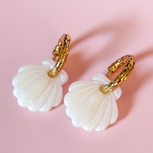 Peut inclure: Une paire de boucles d'oreilles en forme de coquillage. Les boucles d'oreilles présentent un motif de coquillage blanc et scintillant associé à des créoles dorées texturées. Elles sont posées sur un fond rose clair.