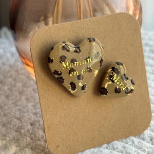 Op de afbeelding: Twee hartvormige oorknopjes in een beige basiskleur met een donkerbruin luipaardpatroon en bladgoudaccenten. Het grotere hart bevat de gouden tekst Maman en or, terwijl het kleinere hart de gouden tekst Mini bevat.