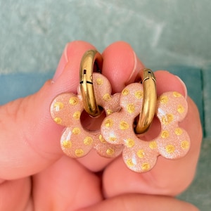 Puede incluir: Un par de pendientes de aro con colgantes en forma de flor. Los colgantes son de color rosa claro con un acabado brillante sutil y están decorados con pequeños lunares de color dorado. Los aros son de metal dorado liso y pulido.