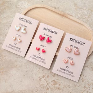 Peut inclure: Trois ensembles de boucles d'oreilles en forme de cœur sur des cartes de présentation rose clair avec les inscriptions Matchy Matchy, Maman et Mini. Le premier est blanc, le deuxième rose vif et le troisième rose pâle. Texte Atelier Bloom Créations.