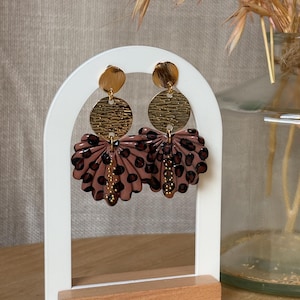 May include: A pair of dangle earrings with a gold-tone textured disc and a fan-shaped drop in shades of brown and black. The earrings are displayed on a white stand with a wooden base, showcasing a unique, artistic design.