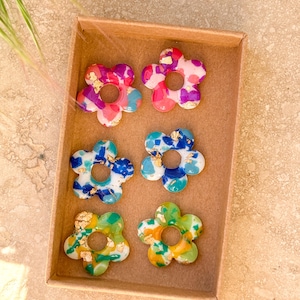 Peut inclure: Six boucles d'oreilles en résine en forme de fleur dans une boîte en carton. Les boucles d'oreilles présentent une variété de couleurs, dont le rose, le bleu, le vert et des accents de feuilles d'or. Chaque boucle d'oreille a un trou au centre.