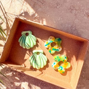 May include: A selection of handmade polymer clay accessories, including two seashell and two flower-shaped pieces. The seashells are light green with ribbed detailing, while the flowers feature yellow, green, and gold accents. The items are displayed in a brown tray.