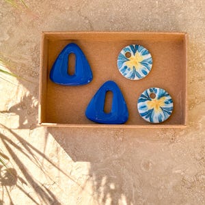 May include: A collection of handmade jewelry components in a small cardboard box. The set includes two cobalt blue triangle-shaped pendants with a central cutout and two round pendants with a blue and yellow floral design.