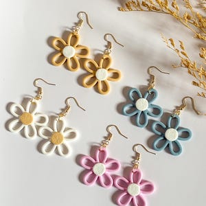 Könnte beinhalten: Sechs Paar ohrringförmige Gänseblümchen in verschiedenen Farben: Gelb, Weiß, Blau und Rosa. Jeder Ohrring hat eine weiße Mitte und goldfarbene Haken. Die Ohrringe sind auf einer weißen Oberfläche angeordnet.