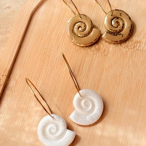 Pode incluir: Dois pares de brincos de argola com pingentes em forma de concha espiral. O par superior é de cor bege brilhante com bordas douradas, enquanto o par inferior é branco cintilante com acabamento em glitter. Cada pingente pende de uma argola dourada.