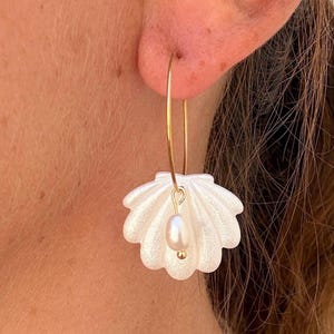 Peut inclure: Une boucle d'oreille créole dorée ornée d'un pendentif en forme de coquillage blanc scintillant. Une petite perle blanche de forme ovale est suspendue au centre du coquillage par un fin fil doré, créant un bijou pendant délicat et superposé.