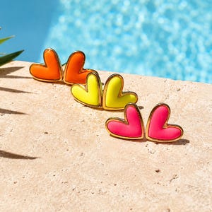May include: A selection of heart-shaped stud earrings in bright colours: orange, yellow, and pink. Each earring has a gold-coloured border. The earrings are displayed on a light-coloured surface with a blue background.
