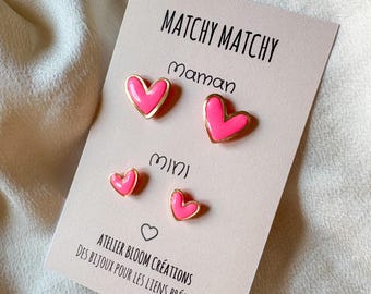Puede incluir: Dos pares de pendientes de botón en forma de corazón rosa neón con bordes dorados montados en una tarjeta de presentación beige. La tarjeta incluye el texto MATCHY MATCHY, Maman, mini y ATELIER BLOOM CRÉATIONS DES BIJOUX POUR LES LIENS PRÉCIEUX.