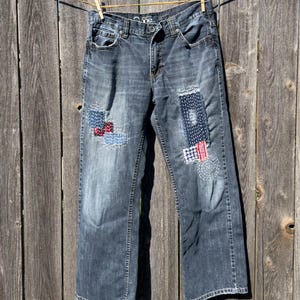 May include: Dark wash denim jeans featuring visible sashiko-style mending with white, red, and blue fabric patches. The jeans include a button fly, belt loops, and five pockets, with decorative white stitching patterns across the front legs.