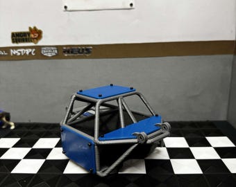 Op de afbeelding: Een blauw en zilveren modelauto-rolkooi staat op een zwart-wit geblokt oppervlak. De kooi heeft een blauwe boven- en voorkant. De achtergrond toont een witte muur met de tekst "fifteen52".