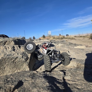 Könnte beinhalten: Ein kleiner, schwarz-silberner ferngesteuerter Rock Crawler klettert einen großen Felsen hinauf. Das Fahrzeug hat große, grobstollige Reifen und einen Überrollkäfig. Der Hintergrund ist ein klarer blauer Himmel und felsiges Gelände.