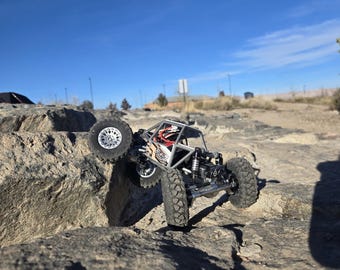 Op de afbeelding: Een kleine, zwart-zilveren op afstand bestuurbare rock crawler beklimt een grote rots. Het voertuig heeft grote, noppenbanden en een rolkooi. De achtergrond is een heldere blauwe lucht en een rotsachtig terrein.