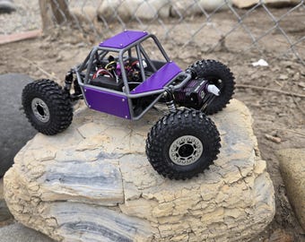 Op de afbeelding: Een paars en grijs op afstand bestuurbare rock crawler speelgoed. Het voertuig heeft grote, zwarte off-road banden en een rolkooi. Het staat op een grote, lichtgekleurde rots. De achtergrond omvat een hek van gaas en een huis.