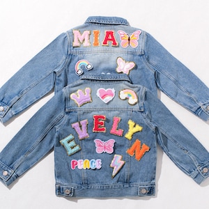 May include: Two light blue denim jackets with colorful patches. One jacket has the name "MIA" and butterfly patches. The other jacket has the name "EVELYN" and patches of a crown, heart, rainbow, and lightning bolt.