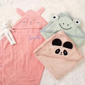 Puede incluir: Tres toallas con capucha para beb&eacute;s en rosa, verde claro y melocot&oacute;n, cada una con un dise&ntilde;o de cara de animal diferente. La toalla rosa tiene orejas de conejo y el nombre "Scarlett" bordado. Tambi&eacute;n hay una peque&ntilde;a mu&ntilde;eca blanca.