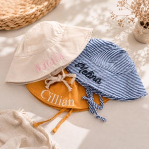 Peut inclure: Trois chapeaux de soleil personnalisés. L'un est beige avec "Natalia" brodé en rose, un autre est jaune moutarde avec "Cillian" brodé en blanc, et le troisième est à rayures bleues et blanches avec "Melina" brodé en noir. Chaque chapeau a une mentonnière.