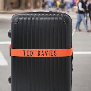 Könnte beinhalten: Schwarzer Rollkoffer mit einem orangefarbenen Gurt mit der Aufschrift "TOD DAVIES". Der Koffer hat ein geriffeltes Design und steht auf einer Straße. Die Räder sind unten sichtbar.