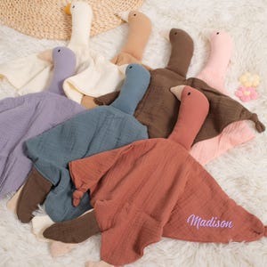 Könnte beinhalten: Eine Sammlung weicher, gänseförmiger Babydecken in verschiedenen Farben, darunter Lavendel, Blau, Braun und Pfirsich. Jede Decke hat einen Gänsekopf mit Schnabel und einen aufgestickten Namen. Die Decken liegen auf einer flauschigen, weißen Oberfläche.