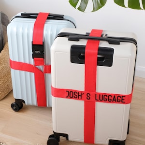 Puede incluir: Dos maletas con ruedas, una azul claro y otra crema, aseguradas con correas rojas brillantes. La maleta crema tiene el texto "JOSH'S LUGGAGE" impreso en la correa. Ambas maletas tienen detalles y ruedas negras.