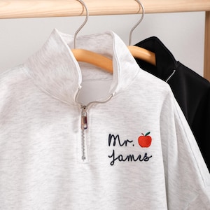 Puede incluir: Una sudadera gris claro con cremallera de cuarto de cierre con el texto bordado "Mr. James" y una manzana roja. La sudadera está colgada en una percha de madera. Una chaqueta negra cuelga en una percha separada en el fondo.