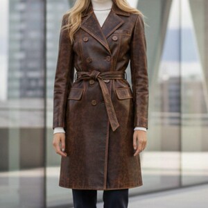 May include: A brown leather trench coat with a double-breasted design and a matching belt. The coat is worn over a white turtleneck and dark blue pants. The model is standing in front of a modern building with glass windows.