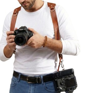 Può includere: Un'imbracatura per fotocamera in pelle marrone con una fotocamera e un obiettivo neri. L'imbracatura è indossata sulle spalle e si collega a una cintura con una seconda fotocamera attaccata. Il fotografo indossa una camicia bianca a maniche lunghe e jeans blu.