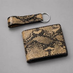 Peut inclure: Un ensemble portefeuille et porte-clés à motif peau de serpent. Le portefeuille est rectangulaire et plié, avec une bordure noire. Le porte-clés est une boucle avec un anneau argenté. Le motif est beige, noir et marron.