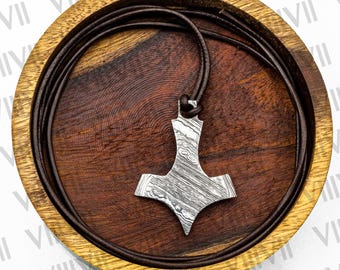 Puede incluir: Un colgante plateado con forma de martillo de Thor, con un diseño estampado, cuelga de un cordón de cuero marrón. El colgante descansa en un cuenco de madera con interior marrón oscuro y borde marrón claro.