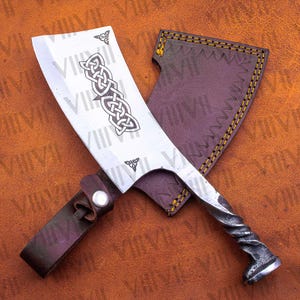 May include: A large, silver cleaver with a Celtic knot design etched on the blade. The handle has a twisted metal design. It is paired with a dark brown leather sheath and strap.