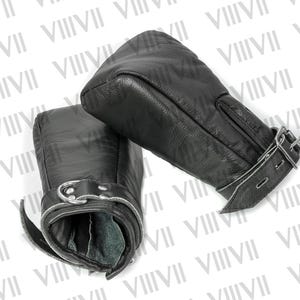 May include: A pair of black leather mittens with a textured surface. Each mitten has a buckle closure with silver-colored hardware. The mittens are designed to cover the entire hand and wrist.